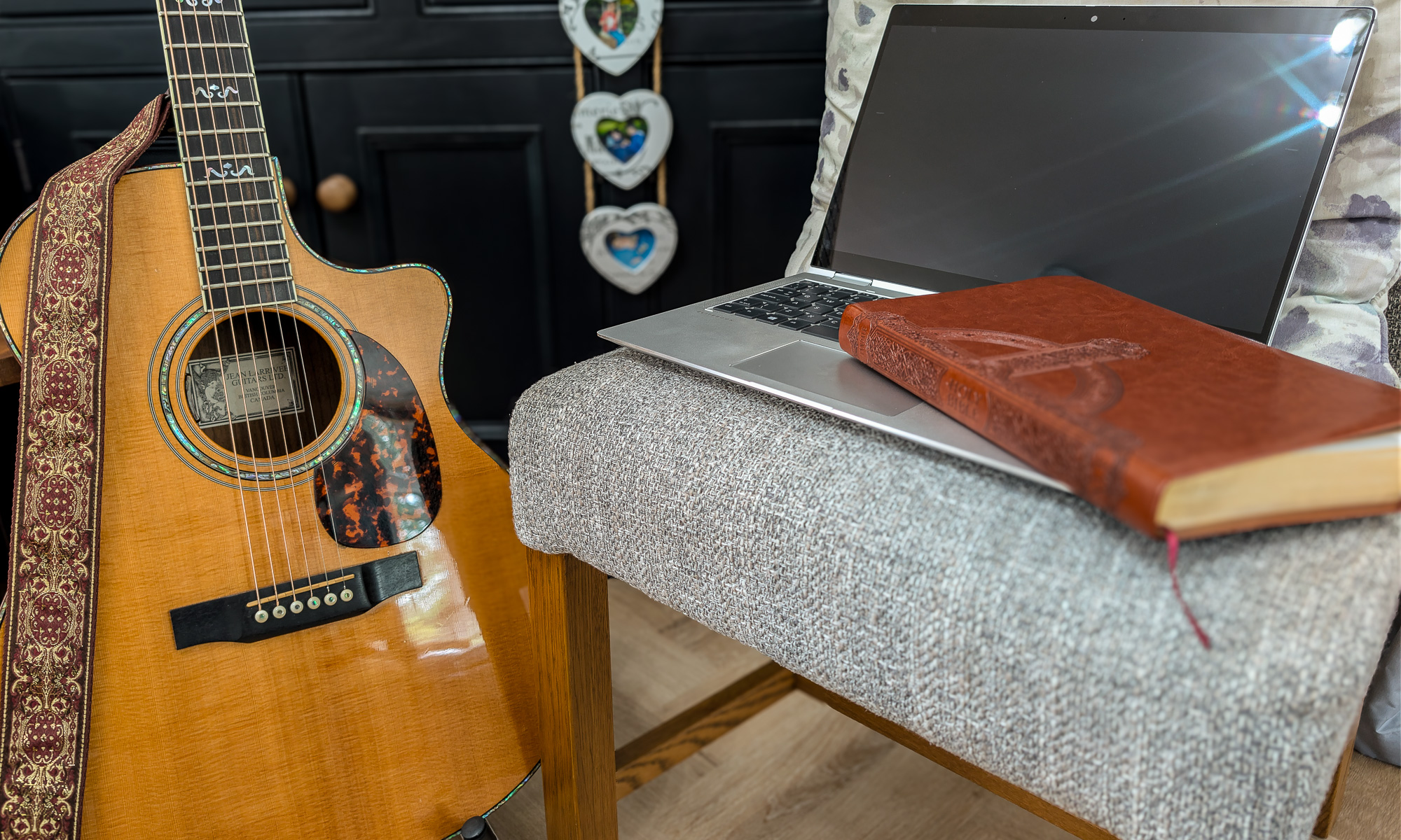 a Larrivée acoustic guitar, laptop and bible in the foreground; a dark sideboard in the background with some photos in heart-shaped frames dangling from a drawer knob